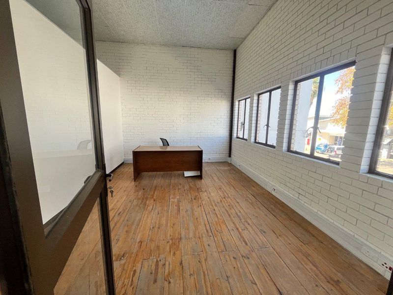 A bright, empty office space featuring exposed white brick walls, timber flooring, and large windows providing natural light.
