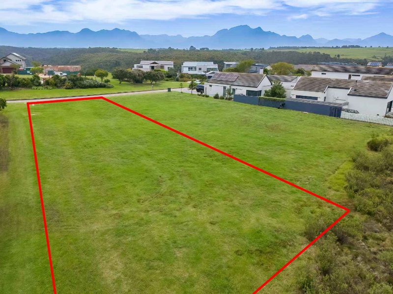 An aerial view of a vacant residential plot featuring level grassed land with surrounding neighborhood views and mountain scenery in the background.