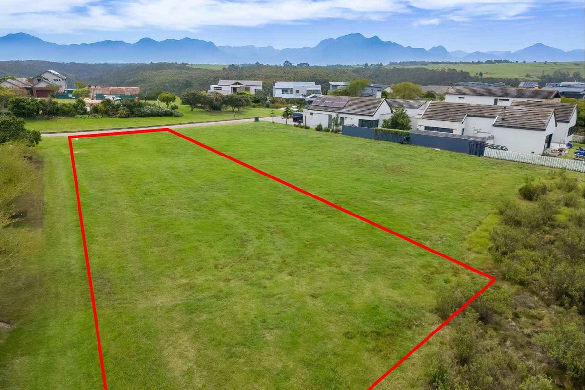 An aerial view of a vacant residential plot featuring level grassed land with surrounding neighborhood views and mountain scenery in the background.