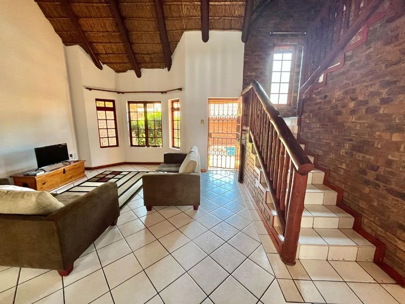 Spacious living area featuring a high thatched ceiling with exposed beams, tiled floors, and a feature exposed brick wall with a wooden staircase.