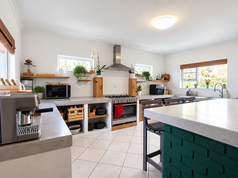 A bright, modern kitchen featuring concrete countertops, a central island with a green brick base, stainless steel appliances, and open timber shelving.