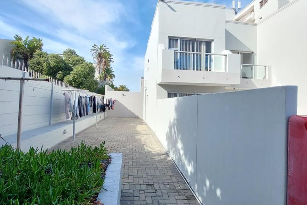A paved rear exterior walkway featuring a retaining wall, security fencing, and a view of the apartment building's upper-level balcony.