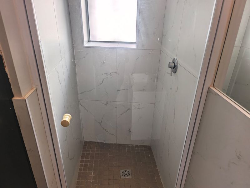 A compact shower stall featuring marble-look wall tiles, mosaic floor tiling, and a modern shower mixer, requiring minor finishing touches to the surrounding wall.