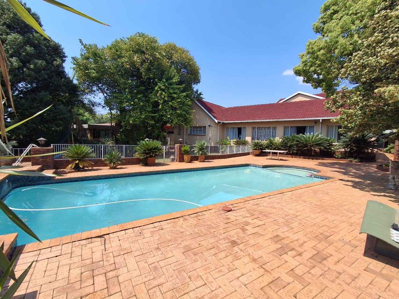 A spacious outdoor pool area featuring a large swimming pool surrounded by brick paving and established garden landscaping.