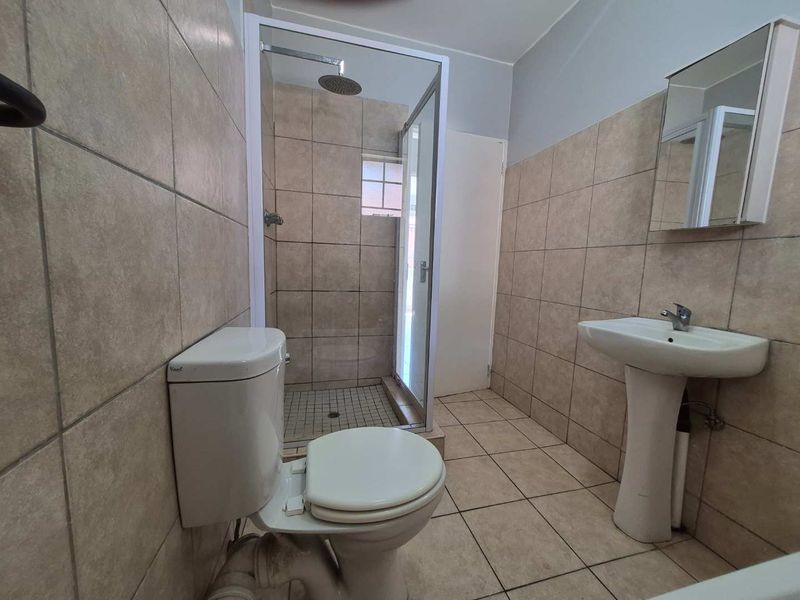 A functional bathroom featuring a pedestal basin, toilet, and a glass-enclosed shower with a rain shower head, finished with neutral wall and floor tiling.