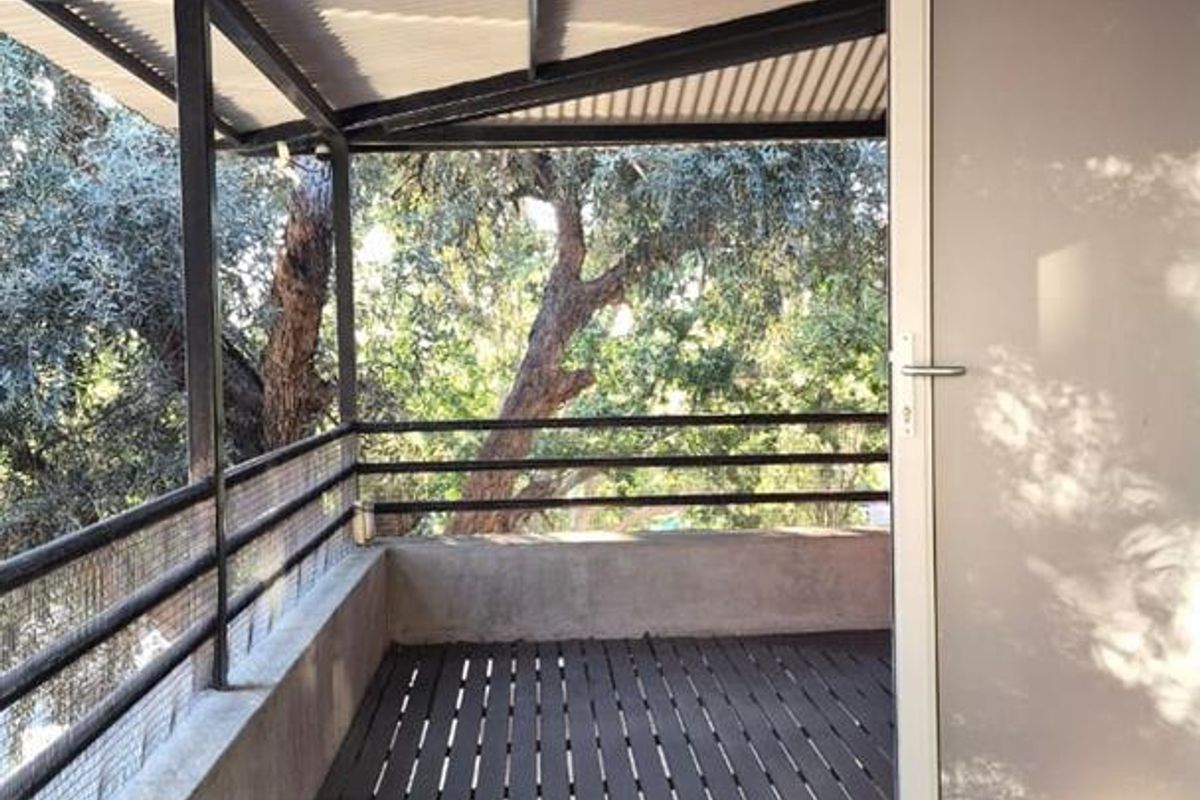 A covered outdoor balcony featuring slatted decking, metal structural framing, and a glass door, offering a peaceful view of surrounding trees.