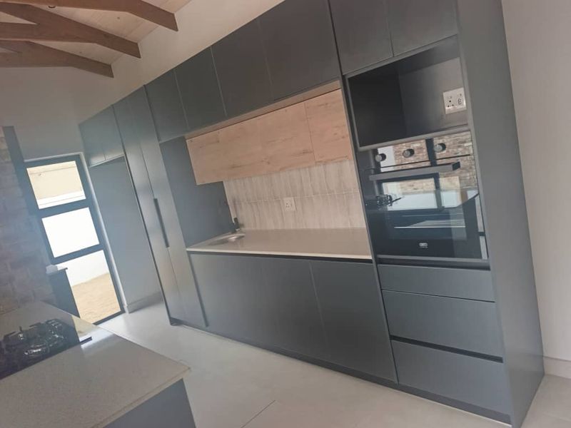Modern kitchen featuring sleek dark cabinetry, light wood accents, stone countertops, integrated oven, gas hob, and exposed timber rafters in a bright, open space.