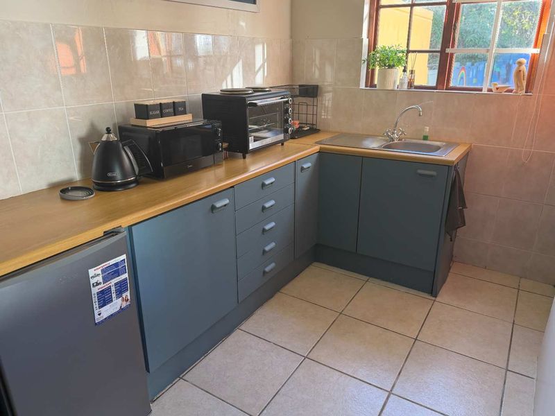 A compact kitchen featuring blue cabinetry, a laminate countertop, stainless steel sink, and tiled splashback with natural light from a window.