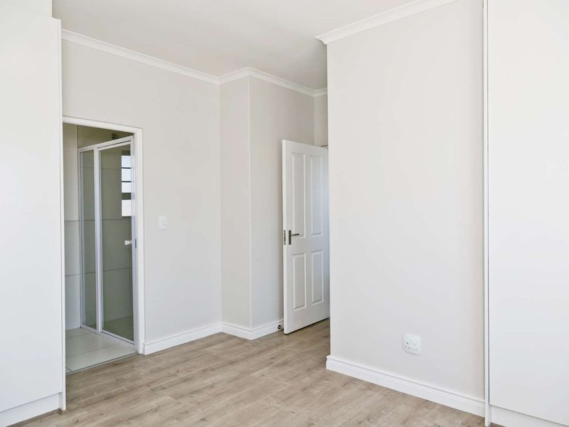 A bright, empty bedroom featuring modern laminate flooring, built-in cupboards, and an attached en-suite shower room with neutral finishes.