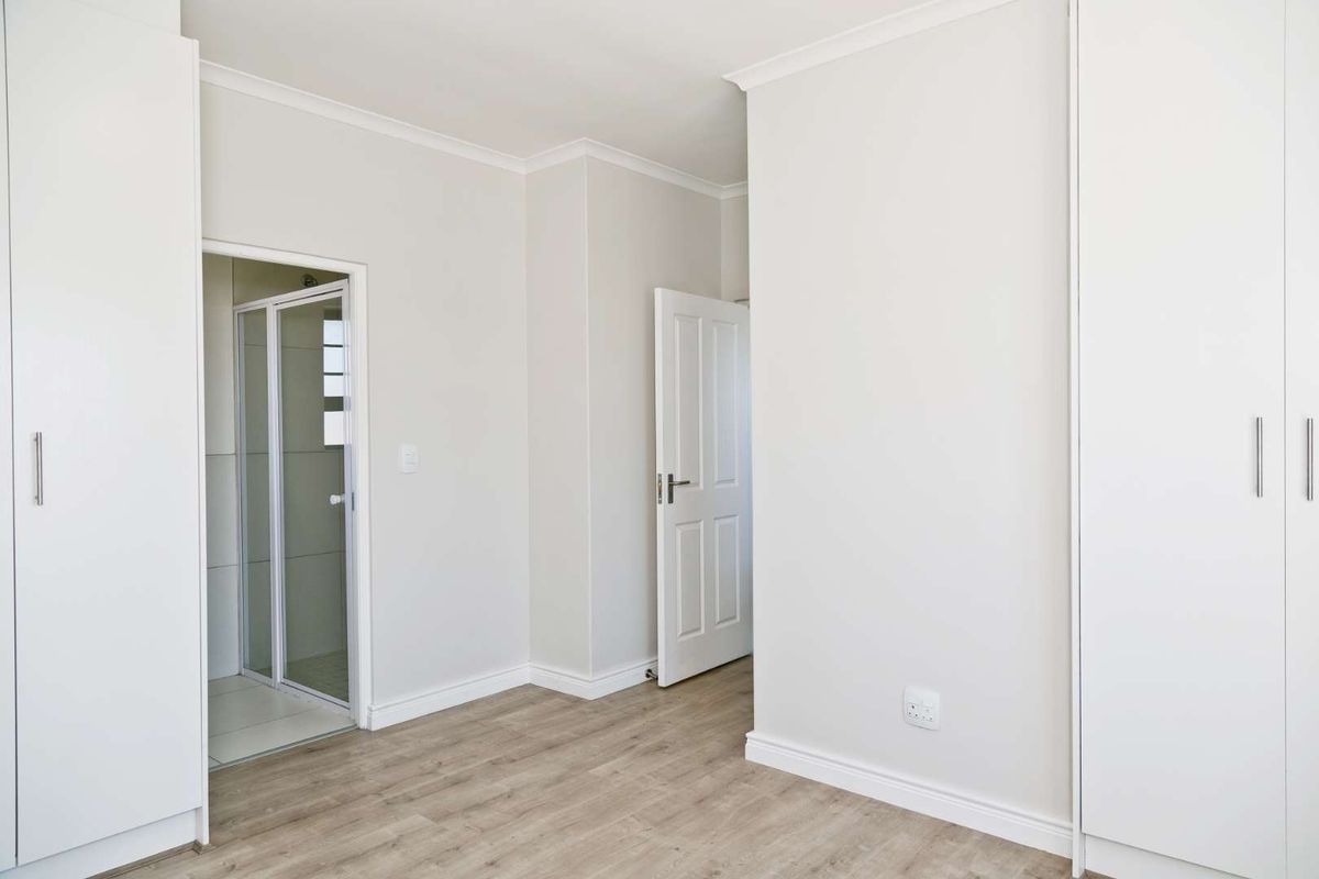 A bright, empty bedroom featuring modern laminate flooring, built-in cupboards, and an attached en-suite shower room with neutral finishes.