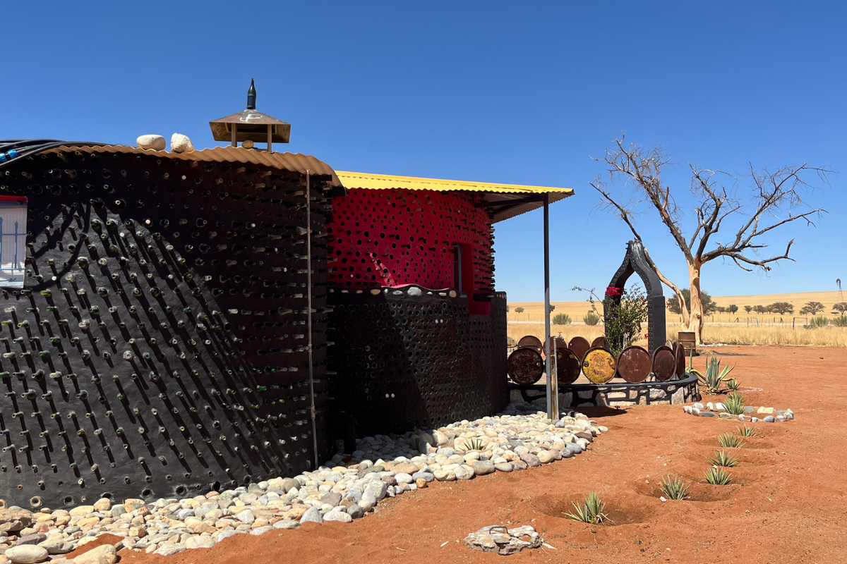 Unique farm dwelling featuring distinctive bottle-wall construction, corrugated roof, and an adjacent outdoor entertainment area set within a vast arid landscape.