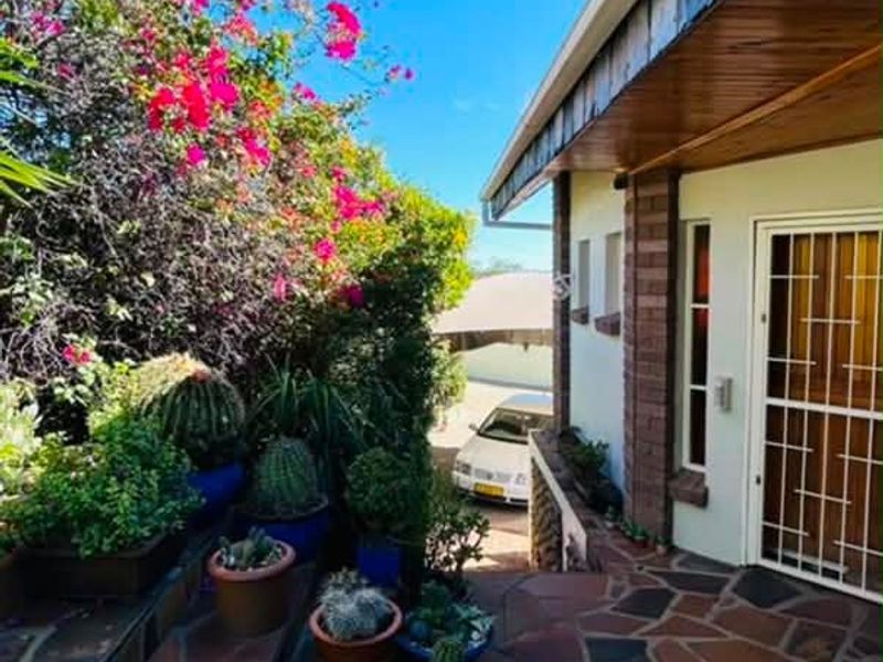 A covered patio area featuring stone paving, timber eaves, and a secure entrance door, surrounded by established garden landscaping.