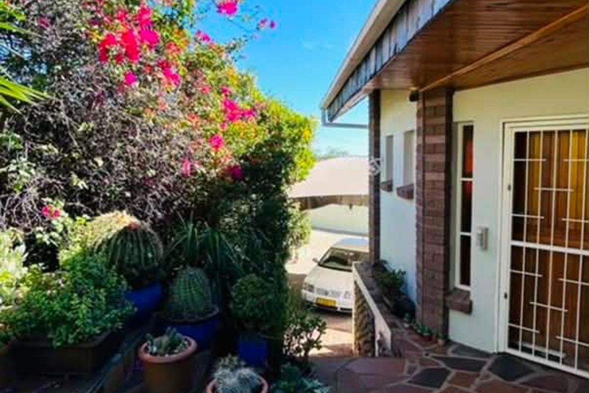 A covered patio area featuring stone paving, timber eaves, and a secure entrance door, surrounded by established garden landscaping.