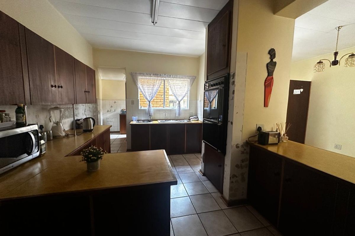 A dated kitchen featuring dark wood cabinetry, laminate countertops, and tiled flooring, with an adjacent scullery area visible through the doorway.