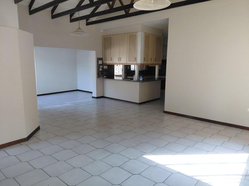 Spacious open-plan living area featuring high vaulted ceilings with exposed timber rafters and neutral tiled flooring, seamlessly connecting to a functional kitchen space.