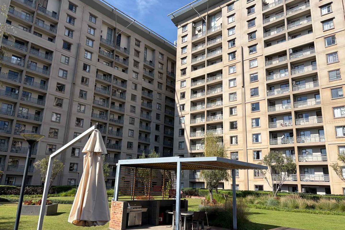 A well-maintained communal garden area featuring a built-in braai station, pergola, and paved patio, set against the backdrop of a modern apartment complex.