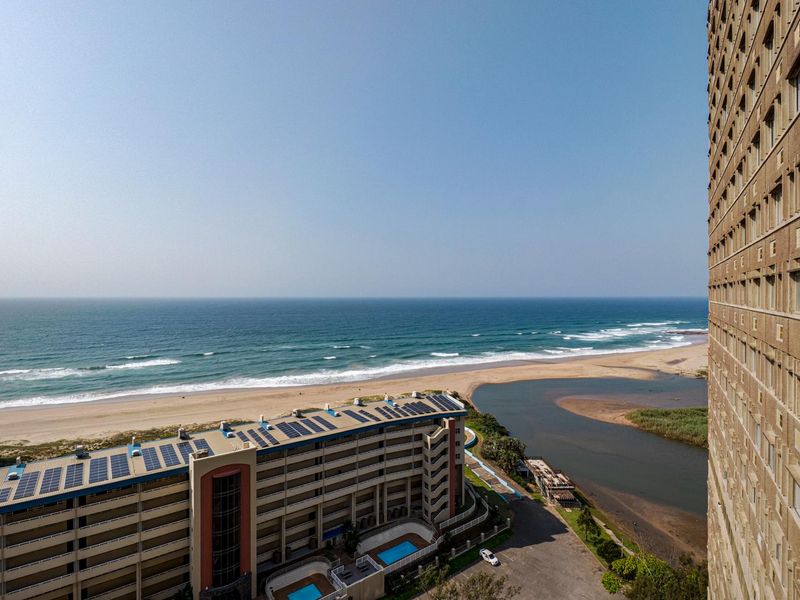 Elevated coastal view overlooking the ocean, sandy beach, and estuary, showcasing the surrounding apartment complex and natural landscape.