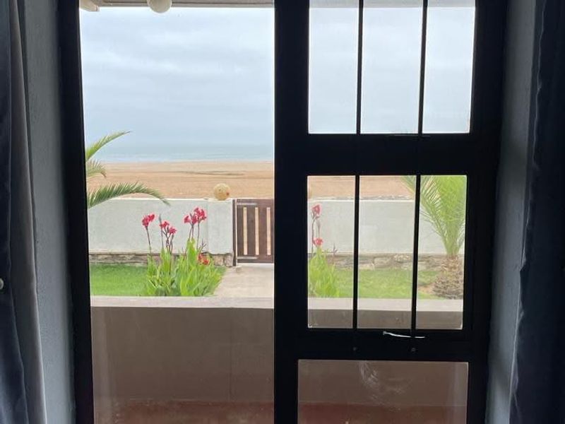 A view through a black metal framed window looking out towards a beach and garden area.