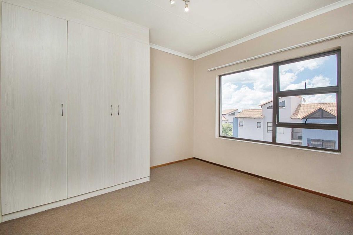 A bright, empty bedroom featuring neutral carpeting, built-in floor-to-ceiling wardrobes, and a large window providing natural light and views of the surrounding complex.
