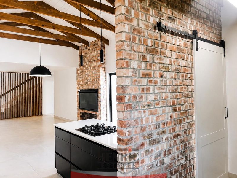 Modern open-plan living area featuring exposed brickwork, timber ceiling beams, a sleek kitchen island with gas hob, and a stylish sliding barn door.