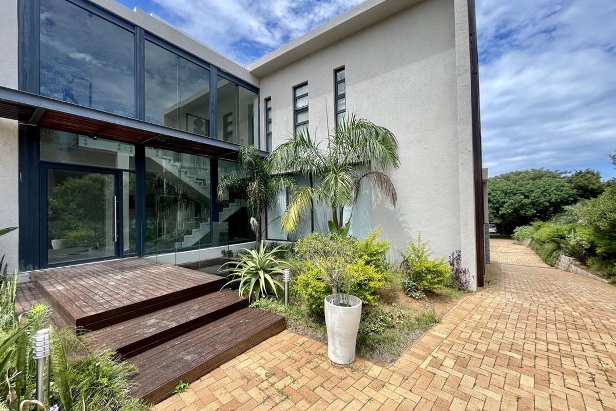 Modern rear exterior featuring a large glass facade, timber decking, and a paved walkway leading through a landscaped garden area.