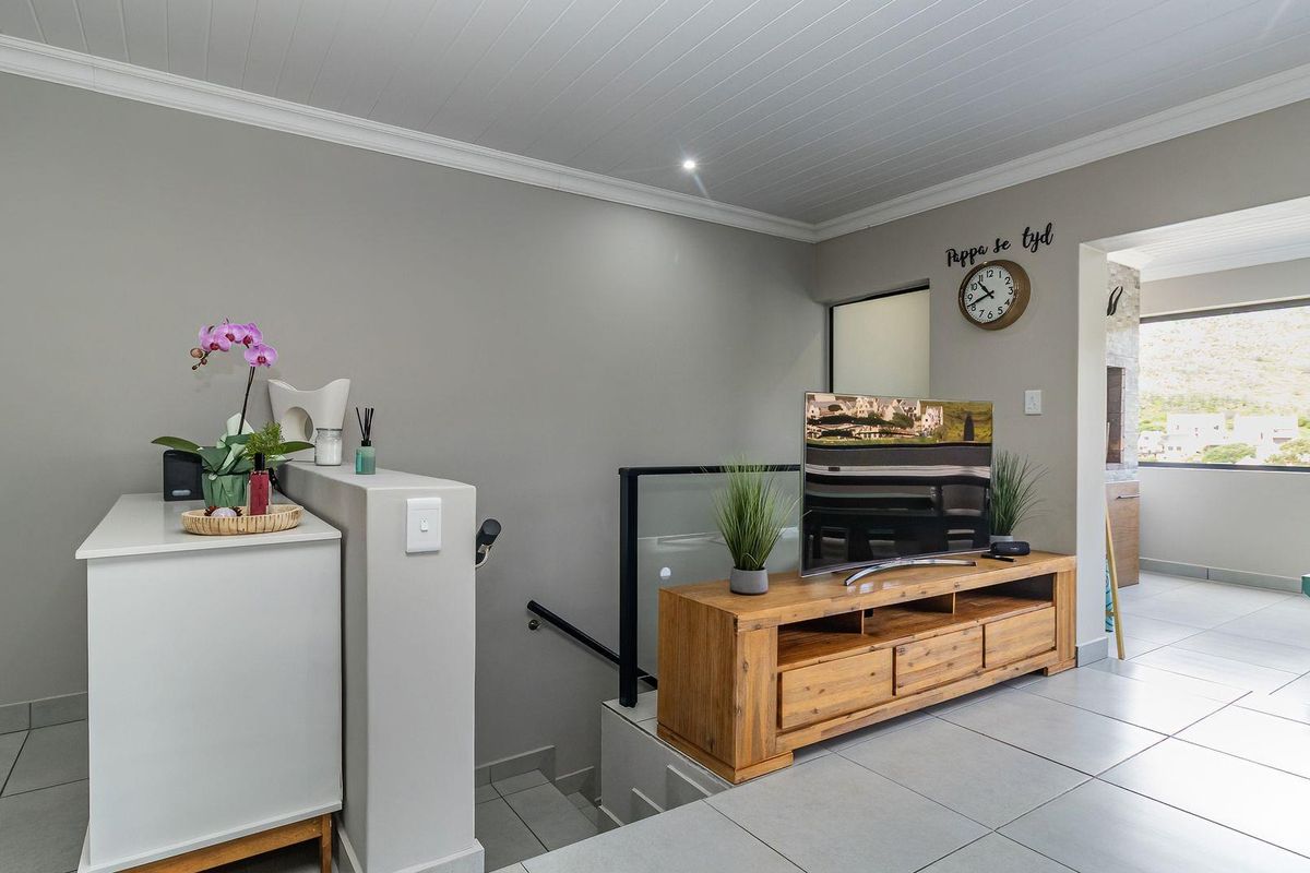 A bright living area featuring tiled floors, a modern glass balustrade staircase, and a clean, neutral aesthetic with recessed ceiling lighting.