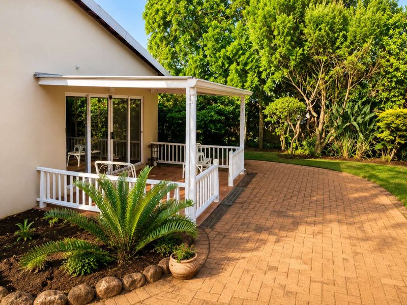 A well-maintained covered patio featuring a white timber balustrade and sliding glass doors, opening onto a paved terrace and lush garden area.