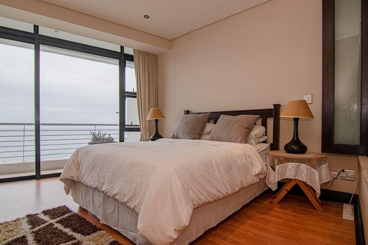 A well-maintained bedroom featuring wood-look flooring, recessed ceiling lighting, and large sliding glass doors opening onto a private balcony with ocean views.