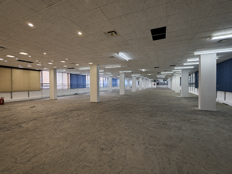 A large, open-plan office space featuring a suspended ceiling, structural columns, and extensive window frontage with vertical blinds.