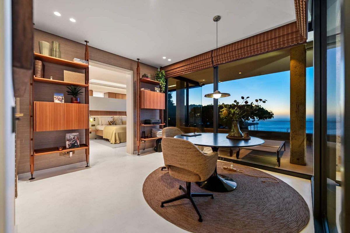 Modern living area featuring floor-to-ceiling glass doors with ocean views, recessed lighting, and custom built-in shelving, seamlessly connecting to the bedroom and balcony.