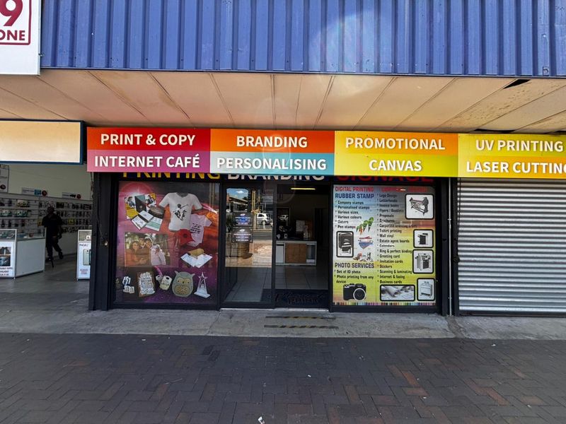 Front facade of a commercial retail unit featuring large glass storefront, prominent business signage, and a metal roller shutter door.