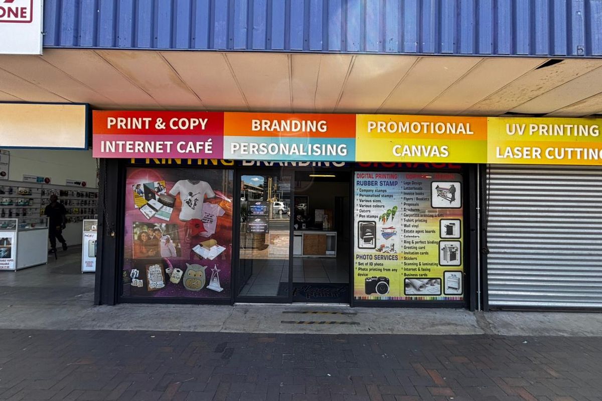 Front facade of a commercial retail unit featuring large glass storefront, prominent business signage, and a metal roller shutter door.