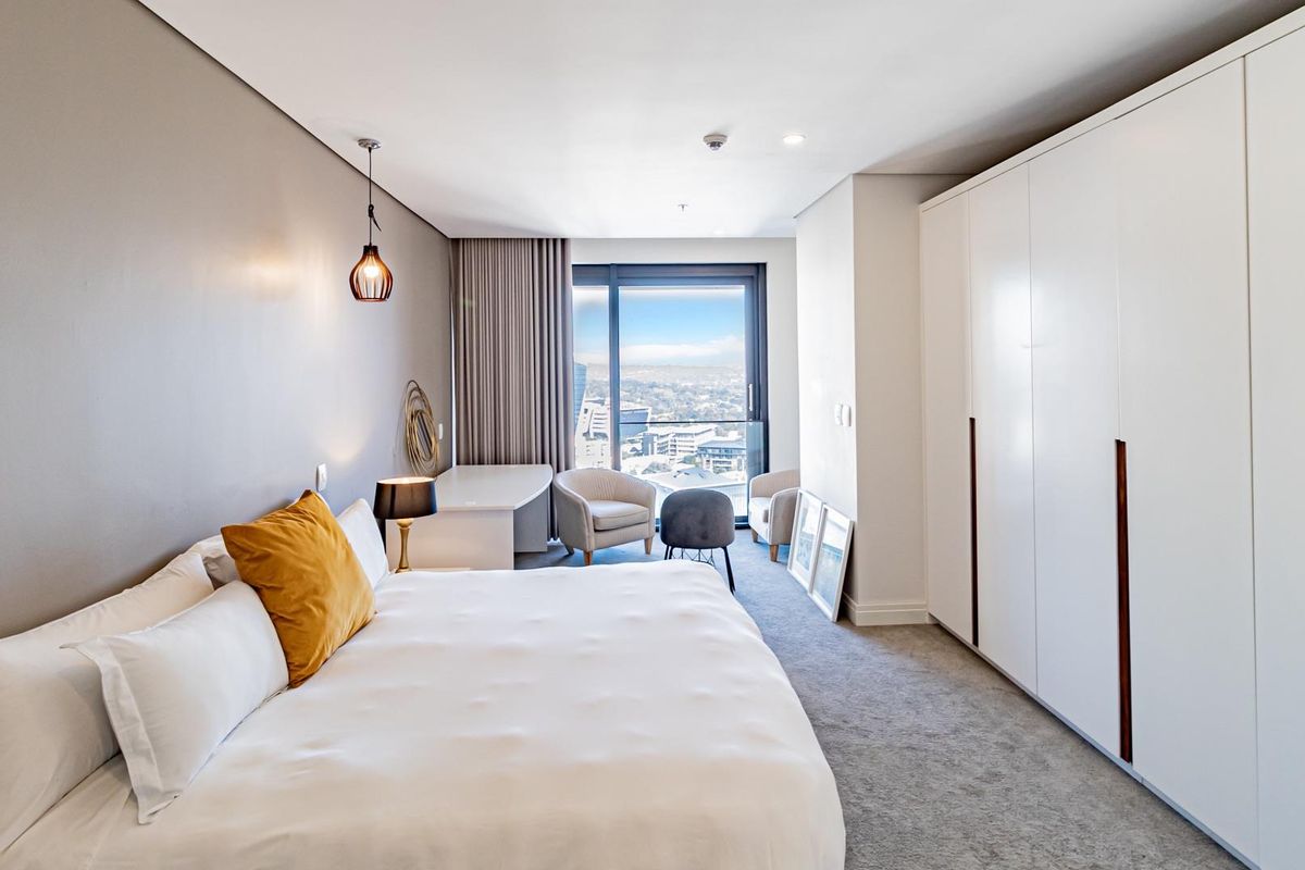 A modern, well-lit bedroom featuring built-in floor-to-ceiling wardrobes, plush carpeting, and large windows offering expansive city views.