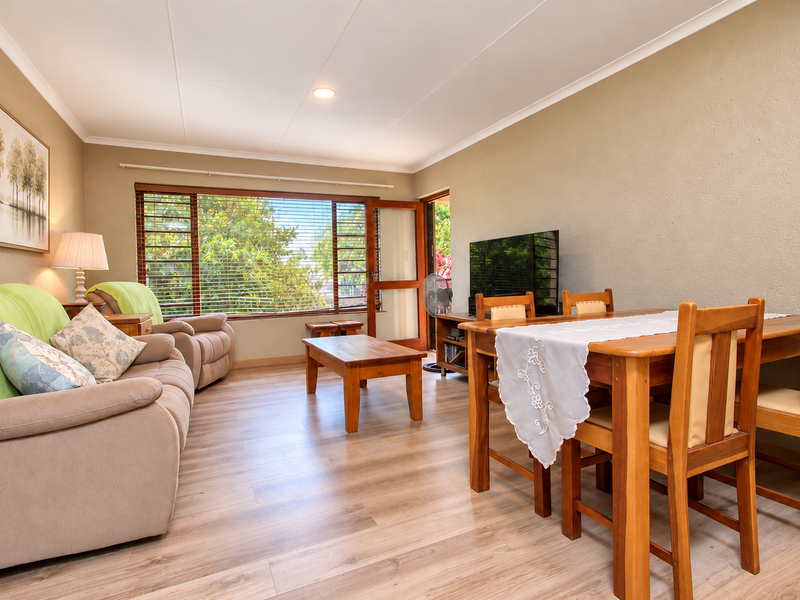 Open-plan living and dining area featuring light-toned laminate flooring, neutral walls, and a large window with venetian blinds providing natural light.