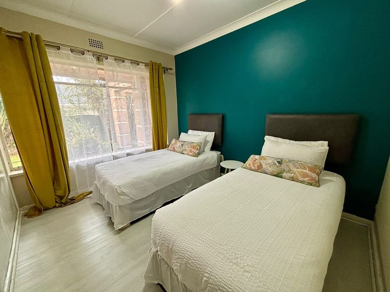 A well-lit bedroom featuring light-toned laminate flooring, a large window with natural light, and a bold teal accent wall.