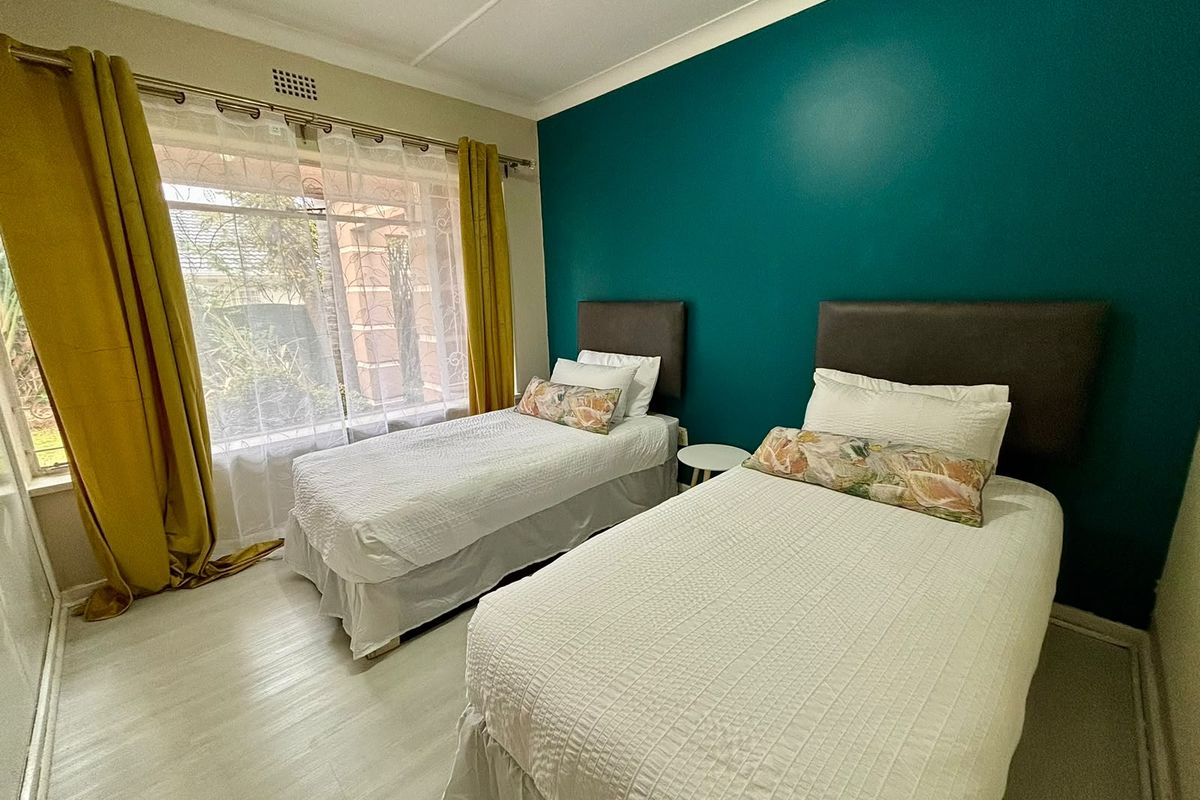 A well-lit bedroom featuring light-toned laminate flooring, a large window with natural light, and a bold teal accent wall.