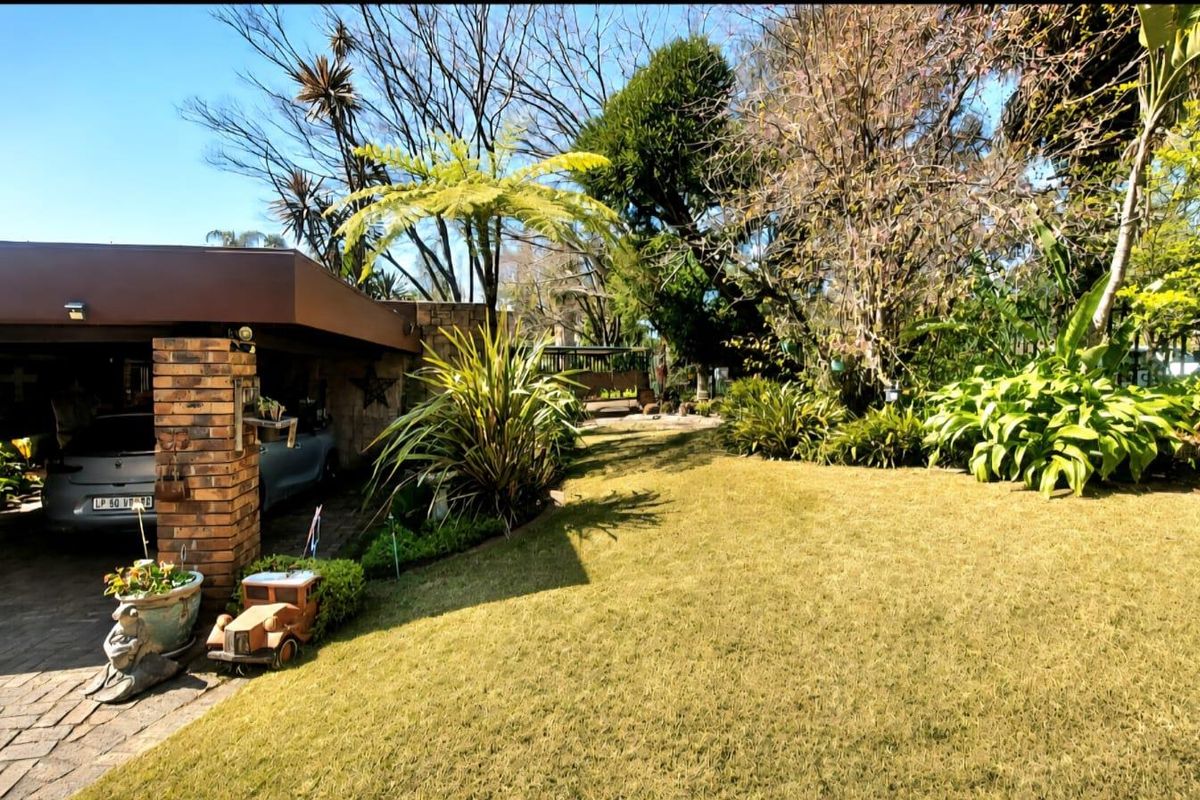A view of the rear garden featuring a paved driveway, a brick-pillared carport, and established landscaping with mature trees and shrubs.