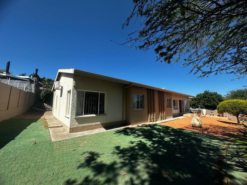 Rear view of the property featuring a paved driveway area, security bars on windows, and established garden space under a clear blue sky.