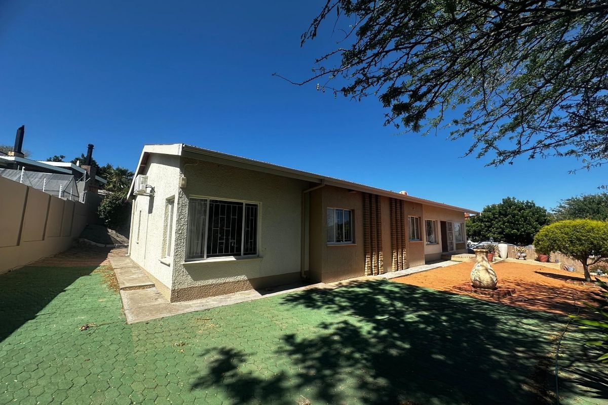 Rear view of the property featuring a paved driveway area, security bars on windows, and established garden space under a clear blue sky.