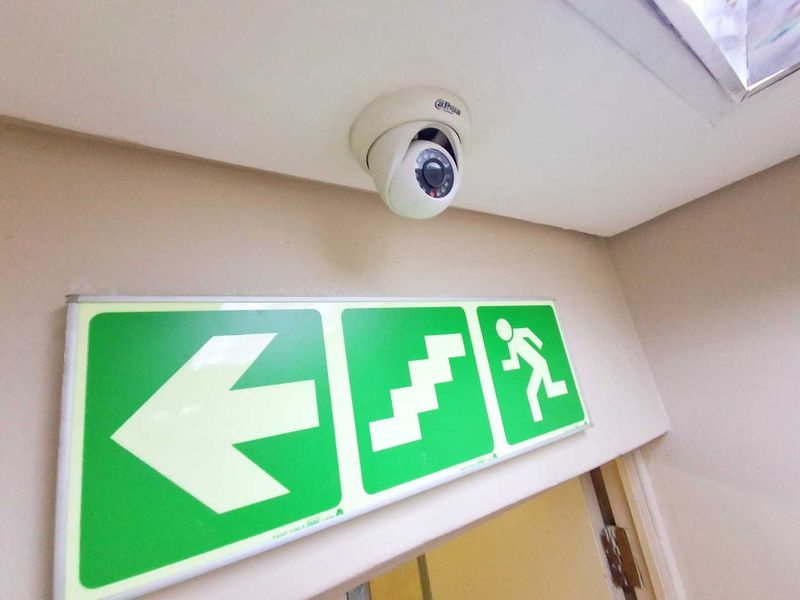 A hallway interior featuring a ceiling-mounted security camera and a prominent green emergency exit sign.