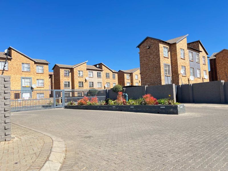 Front view of a multi-story brick apartment complex featuring a paved driveway, secure gated entrance, and landscaped garden beds.