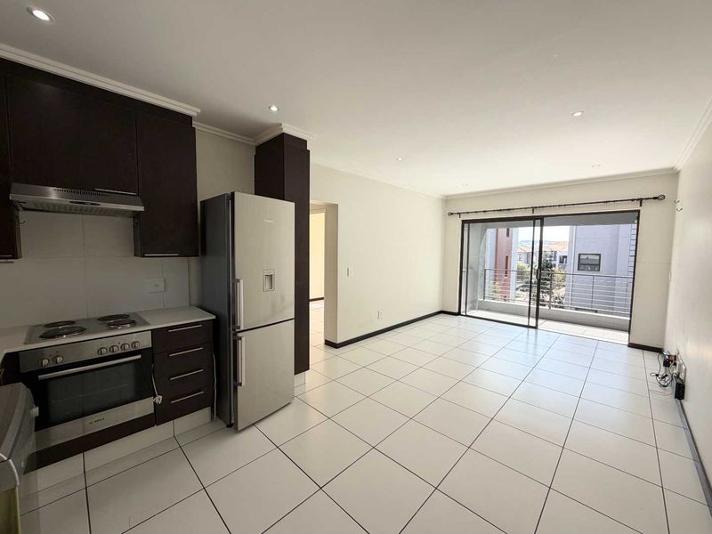 An open-plan kitchen and living area featuring tiled floors, modern dark cabinetry, built-in appliances, and a sliding door leading to a private balcony.