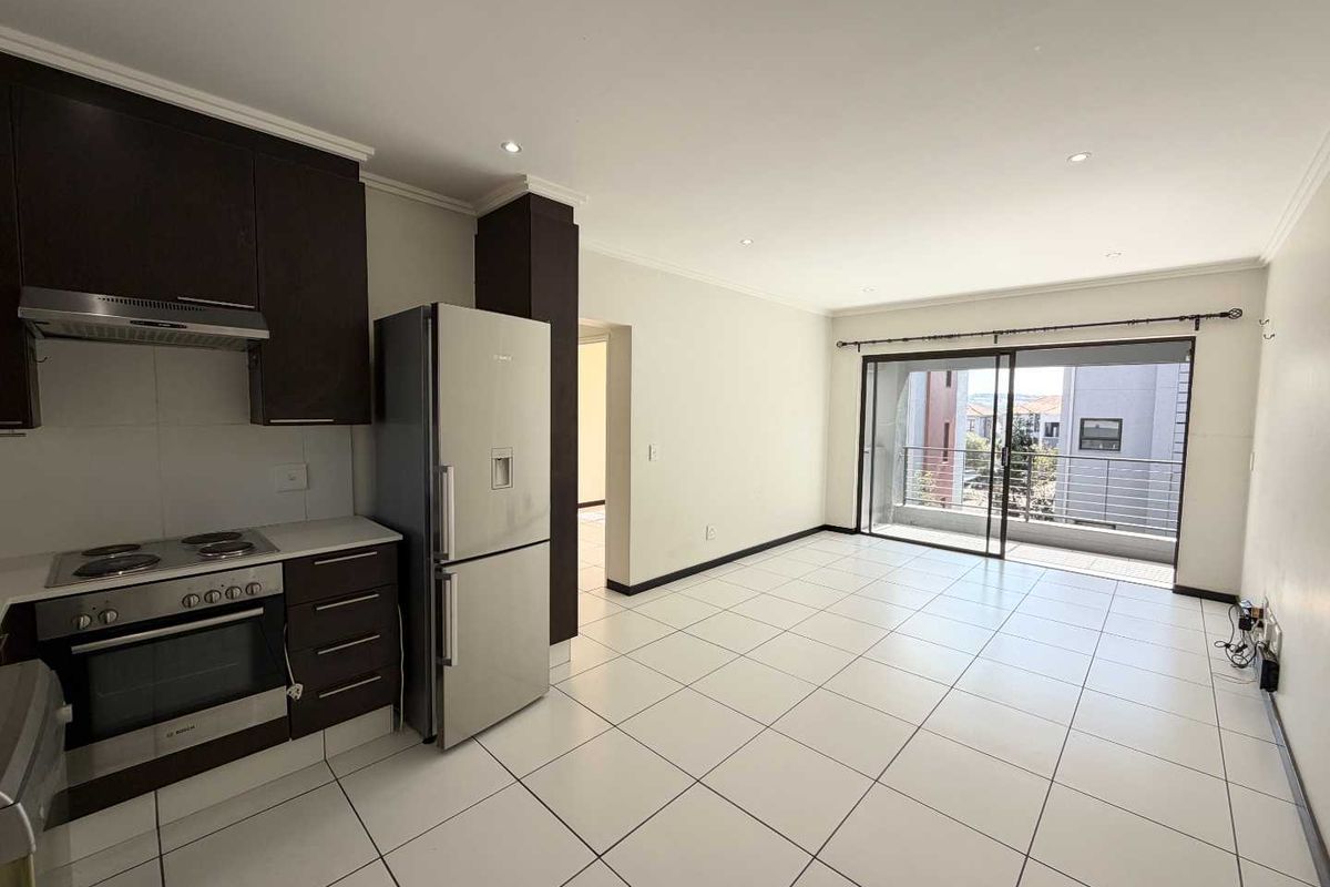 An open-plan kitchen and living area featuring tiled floors, modern dark cabinetry, built-in appliances, and a sliding door leading to a private balcony.