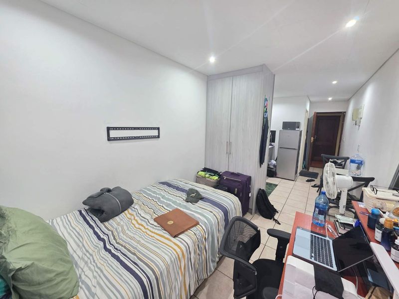 A functional bedroom featuring tiled flooring, recessed downlighting, and a built-in wardrobe, with an adjacent view toward the apartment entrance and kitchen area.