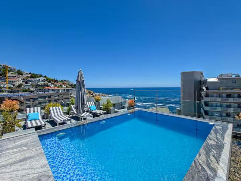 A luxury penthouse pool area featuring a modern swimming pool with stone coping and a glass balustrade, offering panoramic ocean and coastal views.