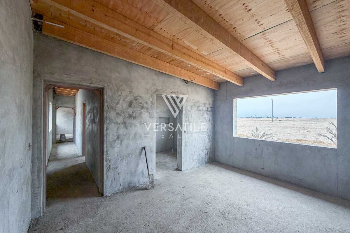 An unfinished living area featuring exposed timber ceiling beams, raw concrete walls, and a large window with an expansive exterior view.
