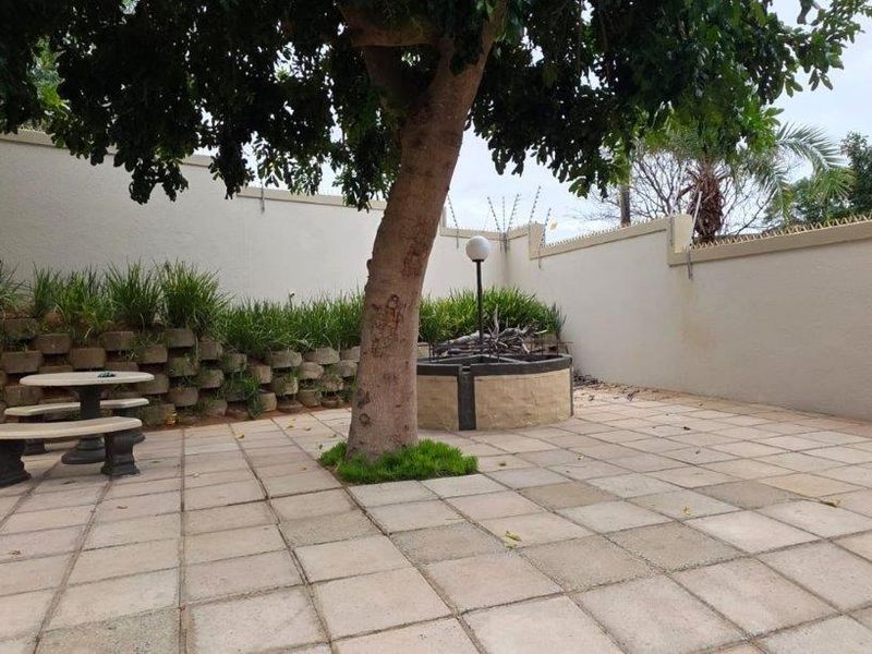 A paved rear courtyard area featuring a retaining wall, perimeter security fencing, and a central garden feature with a mature tree.