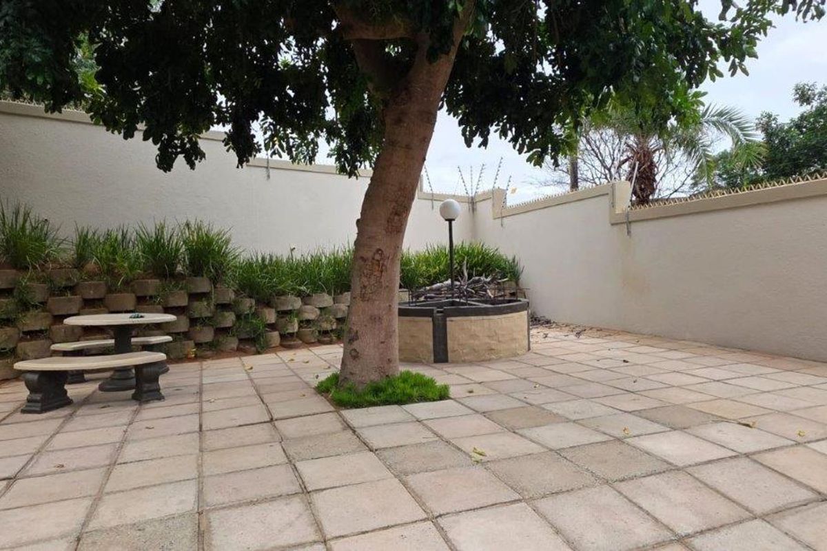 A paved rear courtyard area featuring a retaining wall, perimeter security fencing, and a central garden feature with a mature tree.