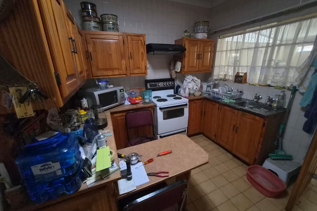 A dated kitchen featuring wooden cabinetry, a freestanding stove with extractor fan, and a stainless steel sink with tiled splashbacks.