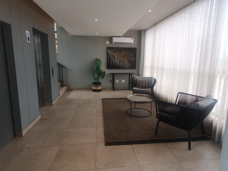 A modern, well-maintained apartment building hallway featuring tiled floors, elevator access, recessed lighting, and large windows providing ample natural light.